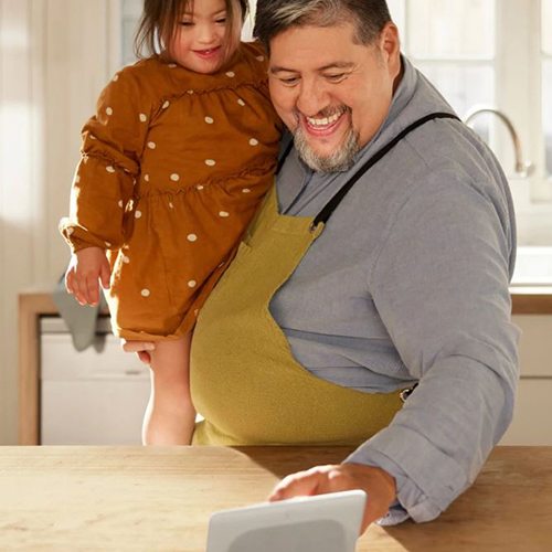 The father holds his toddler daughter in the kitchen and touches the Nest Hub display as they prepare a recipe
