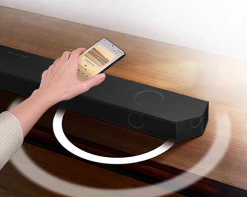 A hand holds a phone playing music and taps it lightly against an Ultra Slim Soundbar. Instantly, the Soundbar gives off a series of round sound waves, indicating that it's playing the same music as the phone.