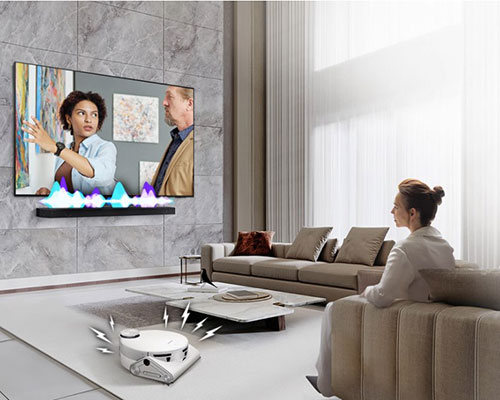 A woman is watching TV, which has an Ultra Slim Soundbar installed directly underneath. The Soundbar has an audio bar indicating the level of volume. A robot vacuum cleaner rolls into the room, giving off distracting noise indicated by lighting bolts. In response, the Soundbar plays at a louder volume, with the audio bar giving off greater activity.