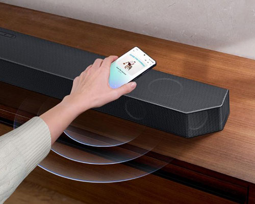 A hand taps a smartphone with the Samsung music app on-screen on the Soundbar. It instantly plays music, showing how easy it is to switch from smartphone to Soundbar.